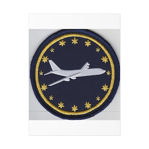 May include: Circular embroidered patch with a navy blue background, a white aeroplane, and gold stars. The design is framed by a gold border. The patch is likely machine-embroidered, with a detailed and precise finish.
