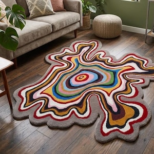 Puede incluir: Una alfombra de forma orgánica e irregular con un patrón concéntrico de colores vibrantes. El diseño consta de bandas onduladas y fluidas en tonos rojo, amarillo, azul, verde, rosa y marrón oscuro, bordeadas por un borde grueso de color gris.