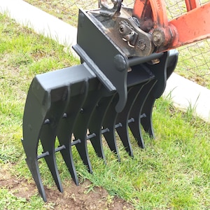 May include: A black grapple attachment for a tractor or excavator. The grapple has multiple curved tines and is attached to an orange and black arm. The image is taken outdoors with green grass in the background.
