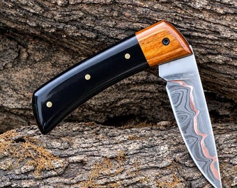 May include: A folding pocket knife featuring a blade with a wavy Damascus steel pattern and a copper-coloured centre stripe. The handle is constructed from a smooth black material and a polished light brown wood section, secured with three brass pins.