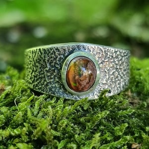 May include: A textured silver ring with a central oval-shaped gemstone. The gemstone displays a mix of brown, orange, and green hues. The ring sits on a bed of green moss, creating a natural and earthy aesthetic.