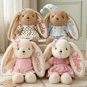 May include: Four plush stuffed bunnies with long ears. The top left bunny is brown with a black and white polka dot dress and the name Jade. The top right bunny is brown with blue pajamas and the name Elena. The bottom left is cream with a pink top and the name Senia. The bottom right is cream with a pink gingham dress and the name Chloe.