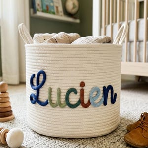 Puede incluir: Una cesta de almacenamiento cilíndrica de cuerda de algodón tejida en color blanco roto con dos asas laterales. El nombre Lucien está escrito en grandes letras cursivas de lana multicolor en tonos azul oscuro, verde salvia, verde bosque, óxido y azul claro.