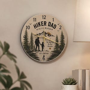 Op de afbeelding: Een ronde wandklok met een crèmekleurige wijzerplaat met de tekst "Hiker Dad Trail Blazer". De wijzerplaat heeft een berg- en boslandschap met een vader en kind die aan het wandelen zijn. De tekst "The Best Dad, The Best Adventure, Est. Forever" is ook opgenomen.