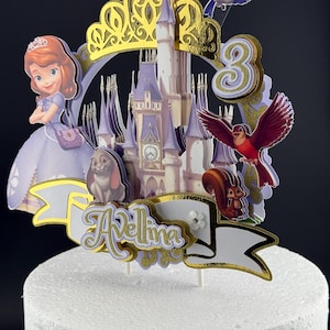 May include: A multi-layered cake topper featuring a cartoon princess in a purple dress, a castle, a rabbit, a squirrel, a blue bird, and a red bird. The design includes a gold crown, the number 3, and a white banner with the name Avellina in gold.