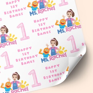 K&ouml;nnte beinhalten: Wei&szlig;es Geschenkpapier mit sich wiederholendem Muster. Das Design enth&auml;lt den Text "Happy 1st Birthday Bambi" und "Ms. Rachel" in Rosa und Blau. Illustrationen einer Frau, eines Monsters und der Zahl 1 sind ebenfalls vorhanden.