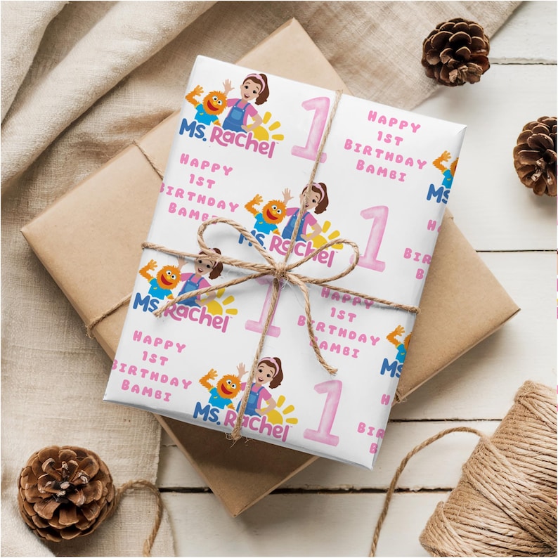 K&ouml;nnte beinhalten: Wei&szlig;es Geschenkpapier mit einem sich wiederholenden Muster mit dem Text "Ms. Rachel", "Happy 1st Birthday Bambi" und der Zahl "1" in Rosa. Das verpackte Geschenk ist mit Bindfaden verschn&uuml;rt.