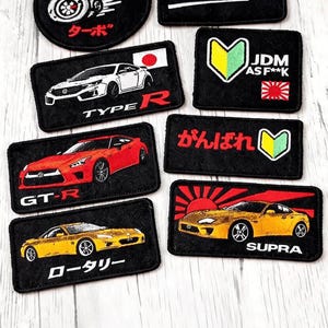 May include: A collection of black embroidered patches featuring Japanese car culture themes. Designs include cars, text like "SEND IT!", "JDM", and Japanese characters. Patches have various colors, including red, white, yellow, and green.