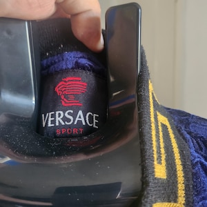 May include: A dark blue velvet jacket featuring a black collar with a gold Greek key pattern. The interior neck label is black with a red Medusa head logo and the white text VERSACE SPORT. The garment is displayed on a black plastic hanger.