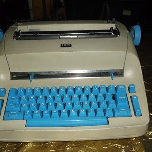 May include: A vintage IBM Selectric typewriter with a light gray body and blue keys. The typewriter has a black carriage return lever and a black ribbon spool.