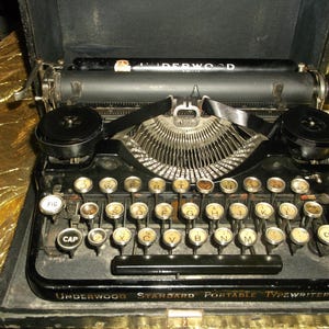 May include: A vintage black Underwood Standard Portable Typewriter in its case. The typewriter features a full keyboard with round, off-white keys and a black ribbon. The word "Underwood" is visible on the top and front of the machine.