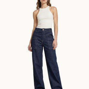 Könnte beinhalten: Eine Frau trägt ein weißes geripptes Tanktop und eine dunkelblaue Jeans mit weitem Bein und großen Vordertaschen. Die Jeans hat eine hohe Taille und wird mit weißen Turnschuhen kombiniert. Das Outfit ist schlicht und lässig.