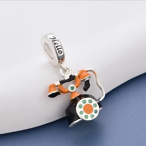Op de afbeelding: Een zilveren bedel in de vorm van een vintage draaischijftelefoon met zwarte en oranje emaille details. De kiesschijf is voorzien van kleine groene kristallen en de bovenste lus is gegraveerd met het woord Hello. De bedel hangt aan een zilveren oogje.