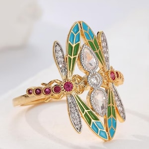 May include: A gold-toned ring featuring two dragonfly motifs with wings accented in blue and green enamel. The design is embellished with small clear stones along the wings and body, complemented by round, deep pink stones set along the ring band.
