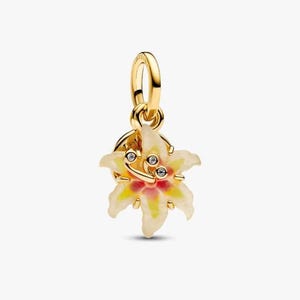 Könnte beinhalten: Ein goldfarbener Anhänger in Form einer Lilienblüte mit cremefarbenen Blütenblättern, einer rosa-gelben Mitte und drei kleinen klaren Steinen im Staubblatt. Der Anhänger verfügt über eine polierte goldene Öse zur Befestigung an einer Kette.