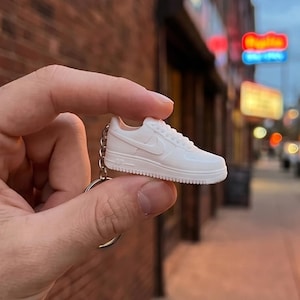 Può includere: Un portachiavi a forma di sneaker bianca in miniatura con un design stampato dettagliato, logo swoosh laterale, lacci frontali e suola testurizzata. La piccola scarpa è fissata a un anello portachiavi in metallo color argento tramite una catena.
