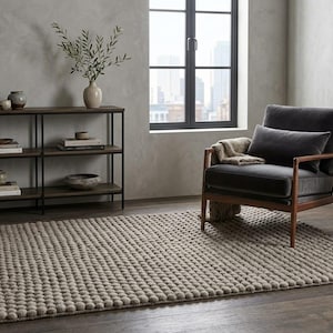 May include: A rectangular area rug with a textured, raised bobble surface in a neutral beige tone. The rug is positioned on a dark wood floor in front of a dark metal shelving unit, a dark gray upholstered armchair, and a small pedestal side table.