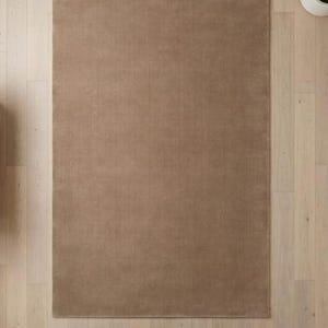 May include: A rectangular, solid brown rug with a soft, textured surface. The rug is placed on a light-colored wooden floor. The rug's neutral color and simple design make it suitable for various interior styles.