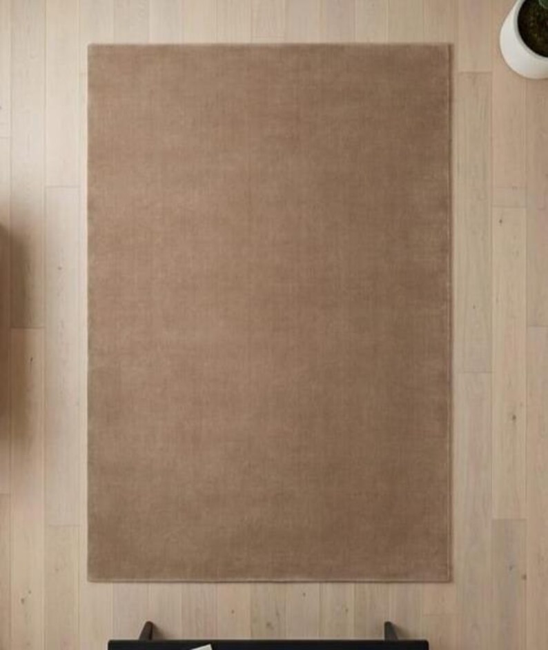 May include: A rectangular, solid brown rug with a soft, textured surface. The rug is placed on a light-colored wooden floor. The rug's neutral color and simple design make it suitable for various interior styles.