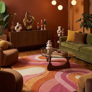 May include: A rectangular area rug featuring a wavy, layered pattern in shades of pink, orange, and cream. The rug is positioned on a wooden floor in a room containing a glass-topped coffee table, a green sofa, two textured armchairs, and a sideboard.