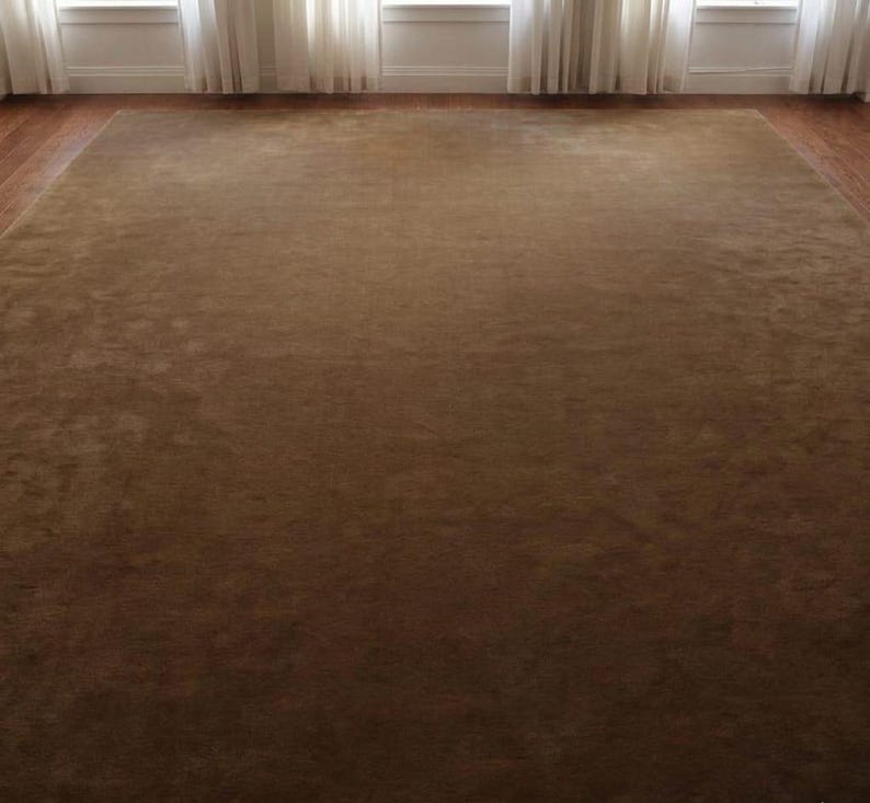May include: A large, rectangular brown rug with a soft, plush texture. The rug fills the majority of the frame, with a dark brown hue. The rug is placed on a hardwood floor, with windows and white curtains in the background.
