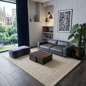 May include: A living room interior featuring a dark gray leather sofa, a low rectangular wooden coffee table, a gray upholstered ottoman, and a large cream-colored textured area rug on dark wood flooring, with a potted plant and wall art.