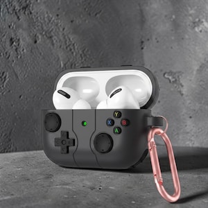 May include: A black protective case for wireless earbuds, designed to resemble a video game controller. The case features a directional pad, buttons, and a green power indicator. The case is open, revealing white earbuds. A rose gold carabiner clip is attached.