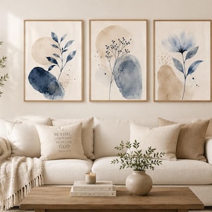 May include: Three framed watercolour art prints with botanical designs in blue, beige, and brown tones. The artwork is displayed above a white sofa with decorative cushions and a wooden coffee table.