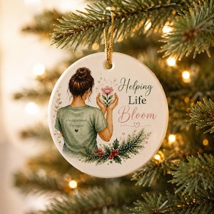 Könnte beinhalten: Ein rundes, weißes Keramikornament mit der Illustration einer Person mit braunem Dutt, die ein grünes T-Shirt mit der Aufschrift Occupational Therapist trägt. Das Motiv zeigt Hände, die eine rosa Blume halten, sowie Stechpalmen, rote Beeren und den Text Helping Life Bloom.
