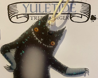 Peut inclure: Ornement de Noël en forme de chat noir patinant sur glace avec des détails scintillants. L'ornement est suspendu à une ficelle blanche et porte le texte "YULETIDE TREE HANGER" sur la carte derrière lui. La carte indique également "HANDMADE WITH LOVE".