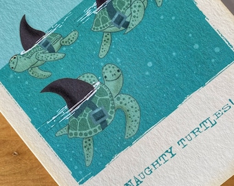 Puede incluir: Una tarjeta de felicitación con un fondo azul que presenta tres tortugas marinas verdes con trajes de aleta de tiburón. El texto "NAUGHTY TURTLES!" está impreso en la tarjeta.