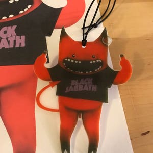 May include: Red cartoon devil with a black t-shirt that says "Black Sabbath" in white letters. The devil is giving the middle finger with both hands. The devil has a tail and is wearing a black string.