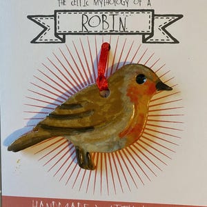 May include: A ceramic robin ornament with a red breast and brown wings, suspended by a red ribbon. The ornament is set against a white background with the text "THE CELTIC MYTHOLOGY OF A ROBIN" and "HANDMADE WITH LOVE".