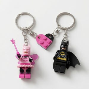 May include: Two keychains featuring Lego minifigures. One is a pink fairy character with wings and a star wand. The other is a black Batman character with a cape. They are connected by a split heart-shaped plastic piece in pink and black.