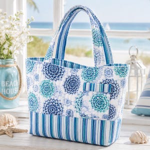 May include: A quilted tote bag featuring a white background with a repeating blue and teal floral mandala pattern. The base and handles are decorated with vertical blue and teal stripes. The bag includes an exterior pocket on the front side.