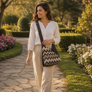 May include: A crossbody bag featuring a prominent black, white, and tan chevron pattern on the main body and a black strap with a repeating circular spiral design. The bag has a structured shape and is worn over the shoulder with a long strap.