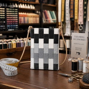 May include: A rectangular crossbody bag featuring a woven pattern of black, white, and gray fabric strips. The bag has a light tan cord strap attached to the top corners. The design consists of vertical columns of alternating colors in a brick-like layout.