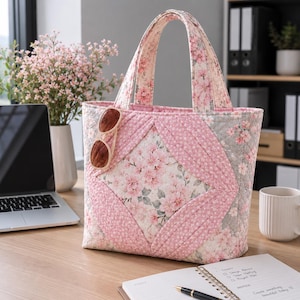 May include: A quilted tote bag featuring a pink and white floral pattern with a central diamond design. The bag has two fabric handles and a pair of brown sunglasses hanging from one side. The exterior is constructed from stitched fabric panels.