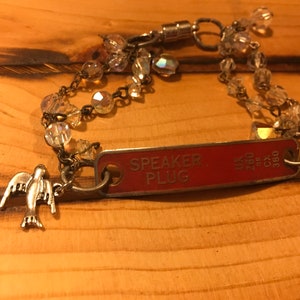 K&ouml;nnte beinhalten: Ein silbernes Armband mit einem roten Metallschild mit der Aufschrift "SPEAKER PLUG" und einem silbernen Taubenanh&auml;nger. Das Armband besteht aus einer Kette und klaren Glasperlen.