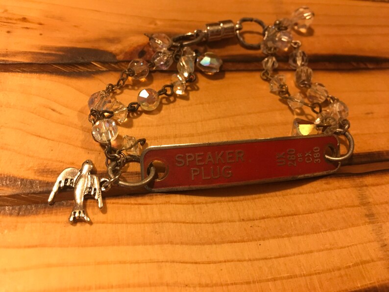 K&ouml;nnte beinhalten: Ein silbernes Armband mit einem roten Metallschild mit der Aufschrift "SPEAKER PLUG" und einem silbernen Taubenanh&auml;nger. Das Armband besteht aus einer Kette und klaren Glasperlen.