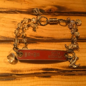 K&ouml;nnte beinhalten: Ein silbernes Kettenarmband mit klaren Kristallperlen, an dem ein rotes Metallschild mit der Aufschrift "SPEAKER PLUG" und "UK 280 CX 380" h&auml;ngt. An dem Schild ist ein silberner Vogelanh&auml;nger befestigt.