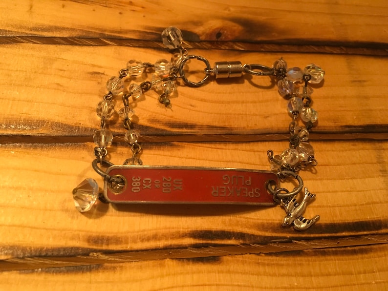 K&ouml;nnte beinhalten: Ein silbernes Kettenarmband mit klaren Kristallperlen, an dem ein rotes Metallschild mit der Aufschrift "SPEAKER PLUG" und "UK 280 CX 380" h&auml;ngt. An dem Schild ist ein silberner Vogelanh&auml;nger befestigt.