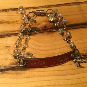 K&ouml;nnte beinhalten: Ein rotes Metallarmband mit einem Anh&auml;nger, auf dem "SPEAKER PLUG" steht, und einem kleinen Vogelanh&auml;nger. Das Armband besteht aus einer Kette mit klaren Kristallperlen und einem Magnetverschluss.