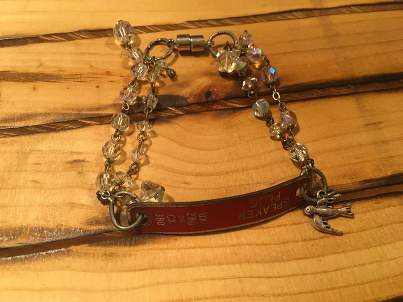 K&ouml;nnte beinhalten: Ein rotes Metallarmband mit einem Anh&auml;nger, auf dem "SPEAKER PLUG" steht, und einem kleinen Vogelanh&auml;nger. Das Armband besteht aus einer Kette mit klaren Kristallperlen und einem Magnetverschluss.