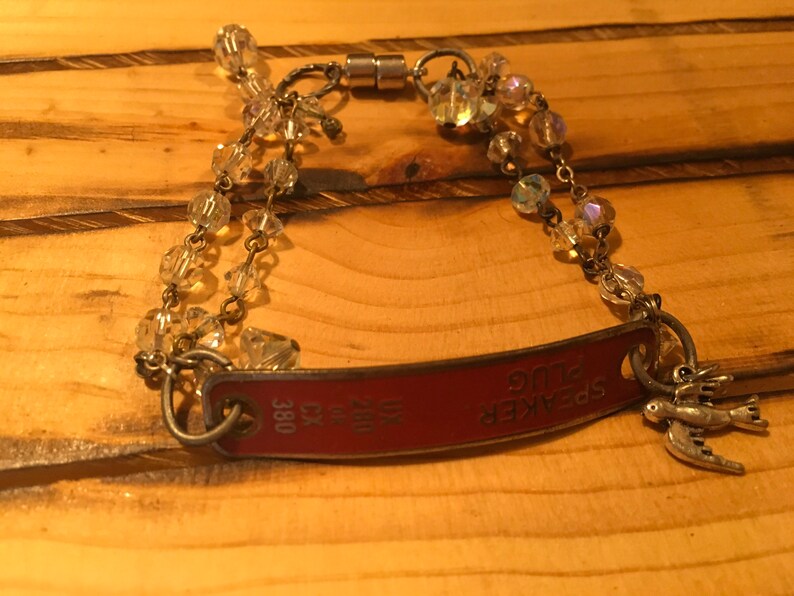 K&ouml;nnte beinhalten: Ein Armband aus klaren Glasperlen und einer silbernen Kette mit einem roten Metallschild, auf dem "SPEAKER PLUG" und die Nummer "380" stehen.