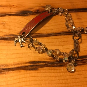 K&ouml;nnte beinhalten: Ein silbernes Armband mit einem roten Metalletikett, auf dem "Speak Please" steht, und einem silbernen Vogel-Anh&auml;nger. Das Armband ist mit klaren Glasperlen besetzt und hat einen Magnetverschluss.