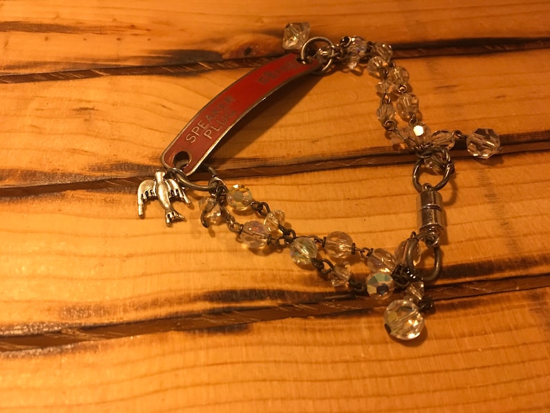 K&ouml;nnte beinhalten: Ein silbernes Armband mit einem roten Metalletikett, auf dem "Speak Please" steht, und einem silbernen Vogel-Anh&auml;nger. Das Armband ist mit klaren Glasperlen besetzt und hat einen Magnetverschluss.