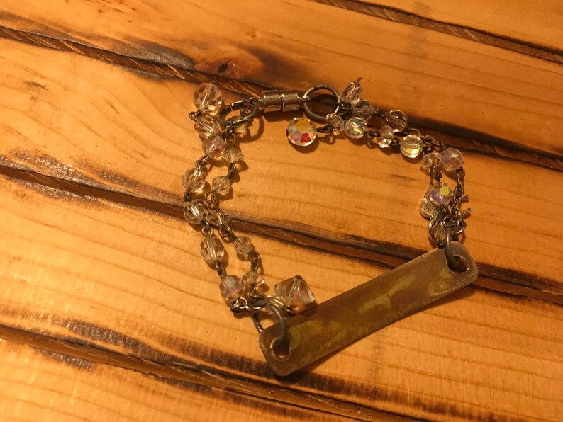 K&ouml;nnte beinhalten: Ein silbernes Armband mit einem rechteckigen Metallschild und Kristallperlen. Das Armband hat einen Magnetverschluss.