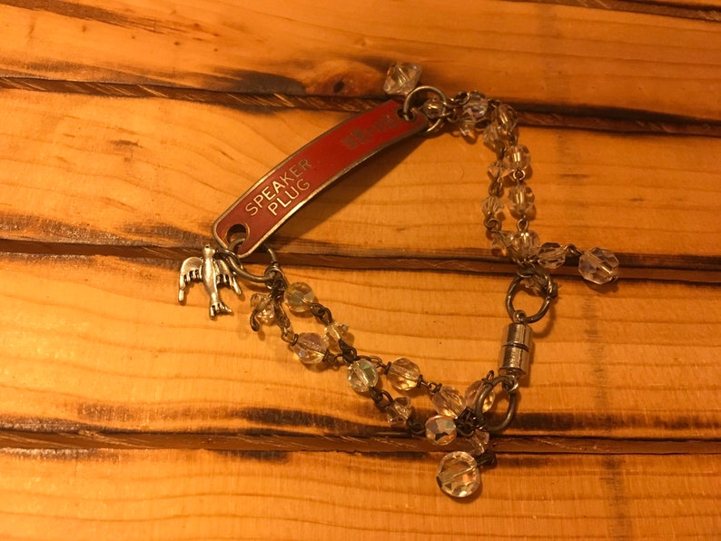 K&ouml;nnte beinhalten: Ein silbernes Armband mit einem roten Metallanh&auml;nger, auf dem "SPEAKER PLUG" steht, und einem kleinen silbernen Vogelanh&auml;nger. Das Armband ist mit klaren Kristallperlen und einem Magnetverschluss versehen.