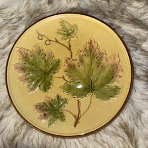 May include: A round ceramic plate with a cream-colored background featuring a raised, embossed design of three green and pink grape leaves on a vine. The plate has a smooth, glossy finish and a dark brown rim encircling the entire edge.